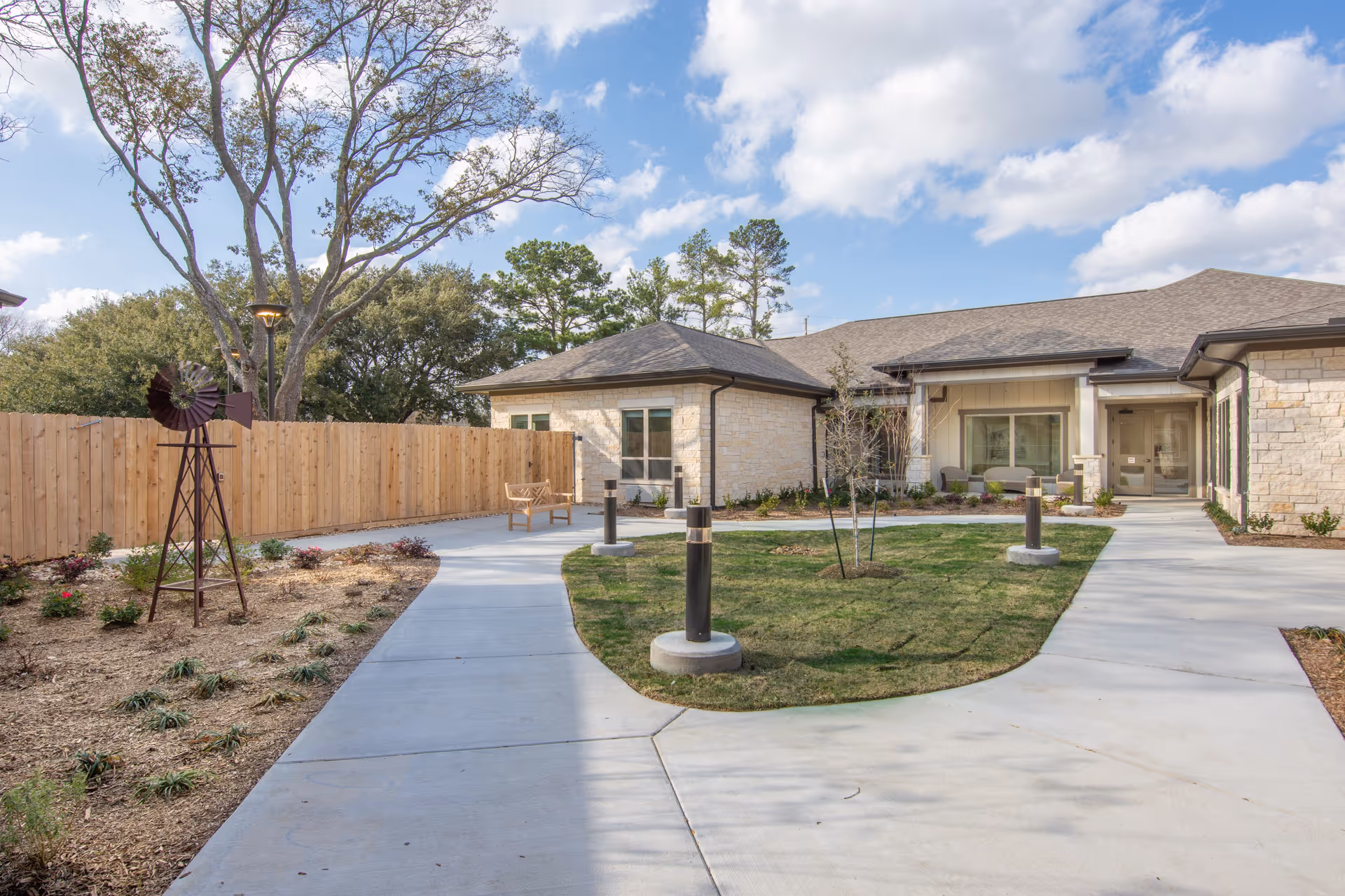 Courtyard with curved concrete paths, lawn, benches and shrubs leading to the stone-clad entrance of a senior living building under a partly cloudy sky.