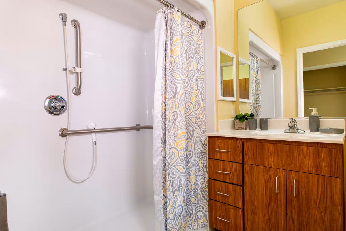 Bathroom with a white shower area featuring a handheld showerhead and grab bars. A patterned shower curtain with gray and yellow designs hangs on a metal rod. Next to the shower is a wooden vanity with a white countertop, a sink, a mirror, and several gray bathroom accessories including a soap dispenser and cups. The walls are painted light yellow.