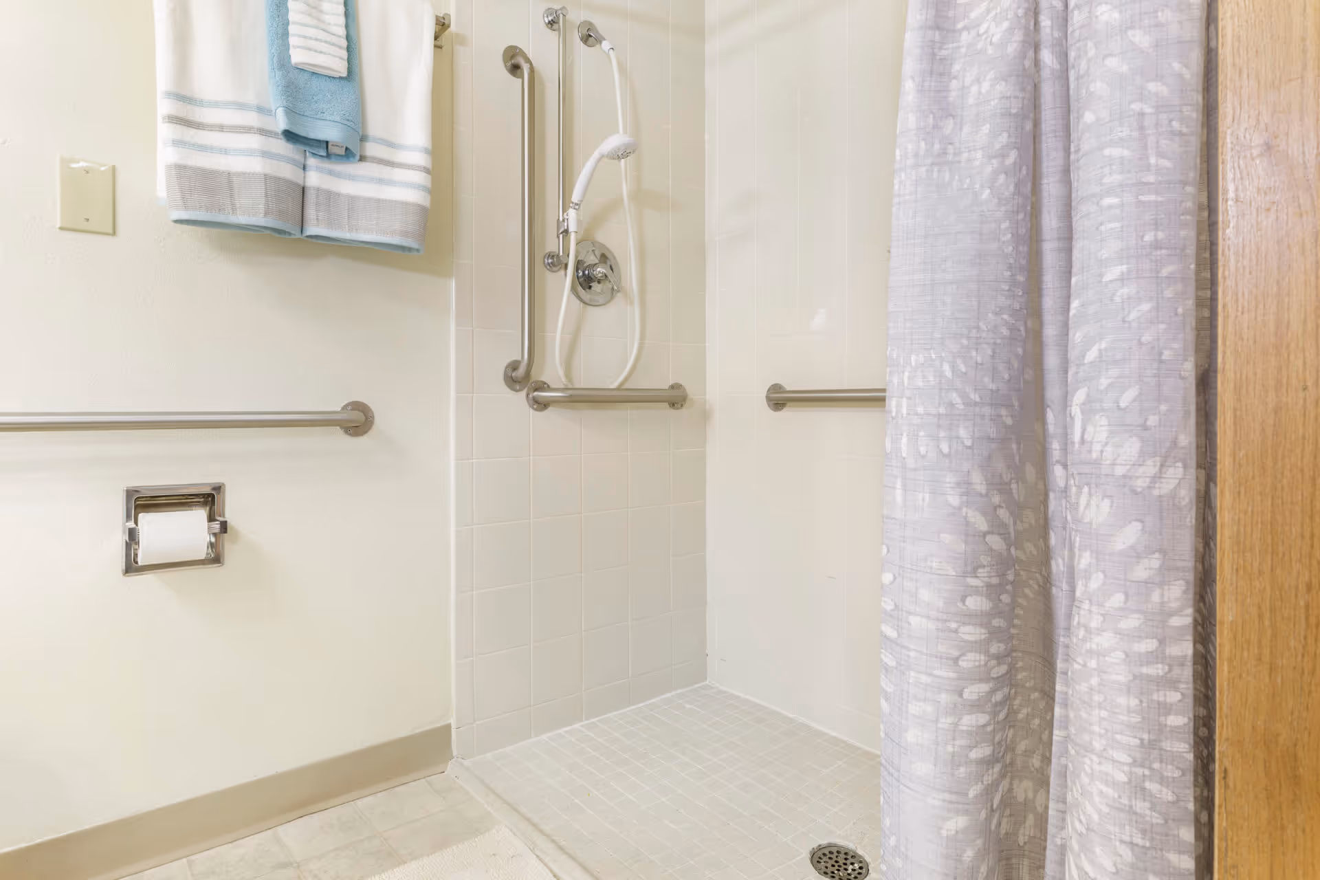 A bathroom shower area with white tiled walls and floor, equipped with multiple stainless steel grab bars and a handheld showerhead. A gray and white patterned shower curtain is partially drawn to the right. On the left wall, there are two folded towels hanging on a towel rack above a toilet paper holder.
