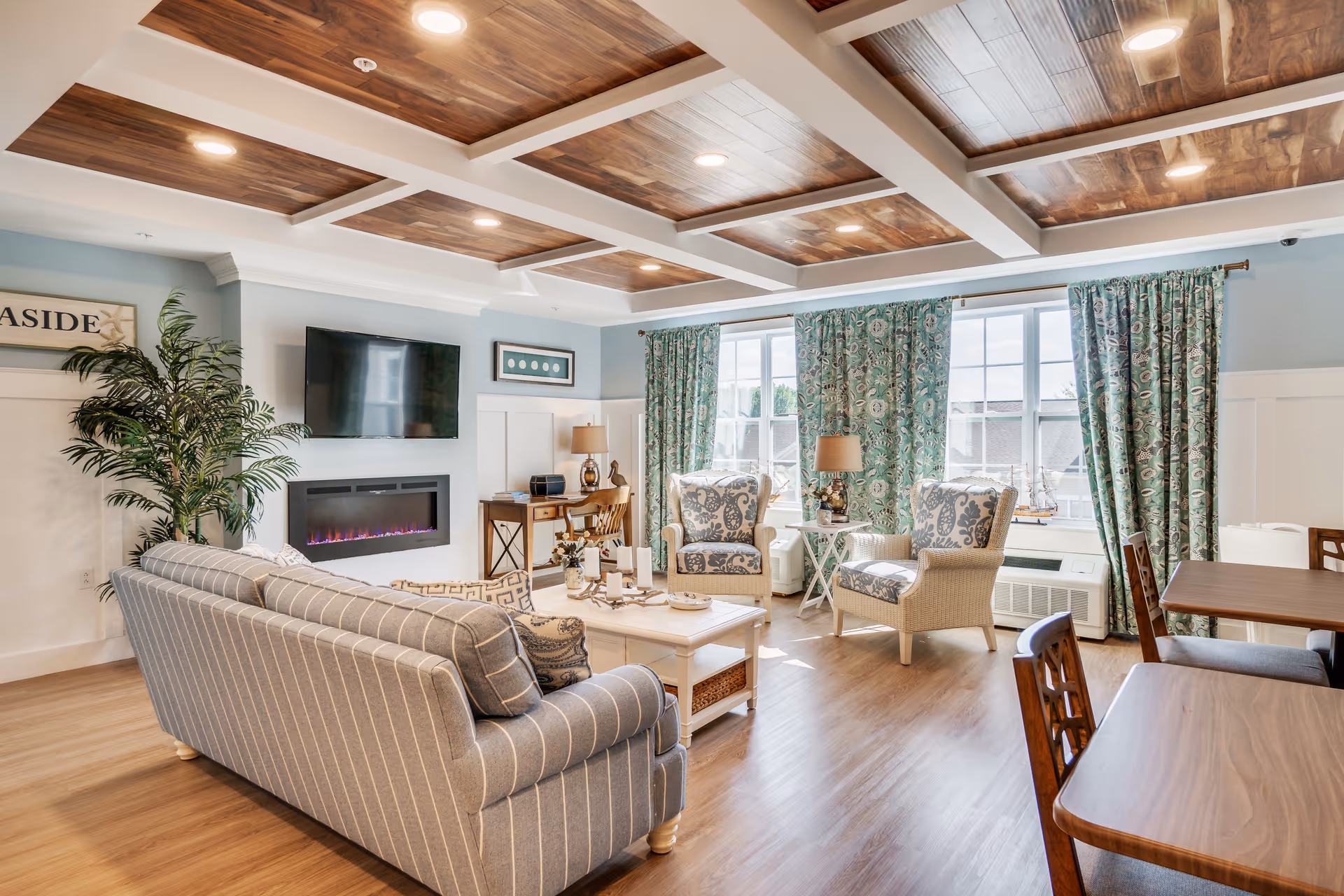 Sunlit common living room with a striped sofa, patterned armchairs, coffee table, wall-mounted TV above an electric fireplace, and large windows with patterned curtains.