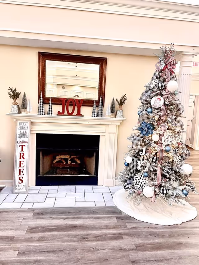 A cozy indoor scene featuring a decorated Christmas tree with ornaments, ribbons, and snowflake decorations next to a white fireplace mantel. The mantel has small decorative trees, a large mirror above it, and red letters spelling 'JOY'. A vertical sign next to the fireplace reads 'Farm Fresh Christmas Trees Cut & Carry'. The floor is wood with a tiled hearth in front of the fireplace.