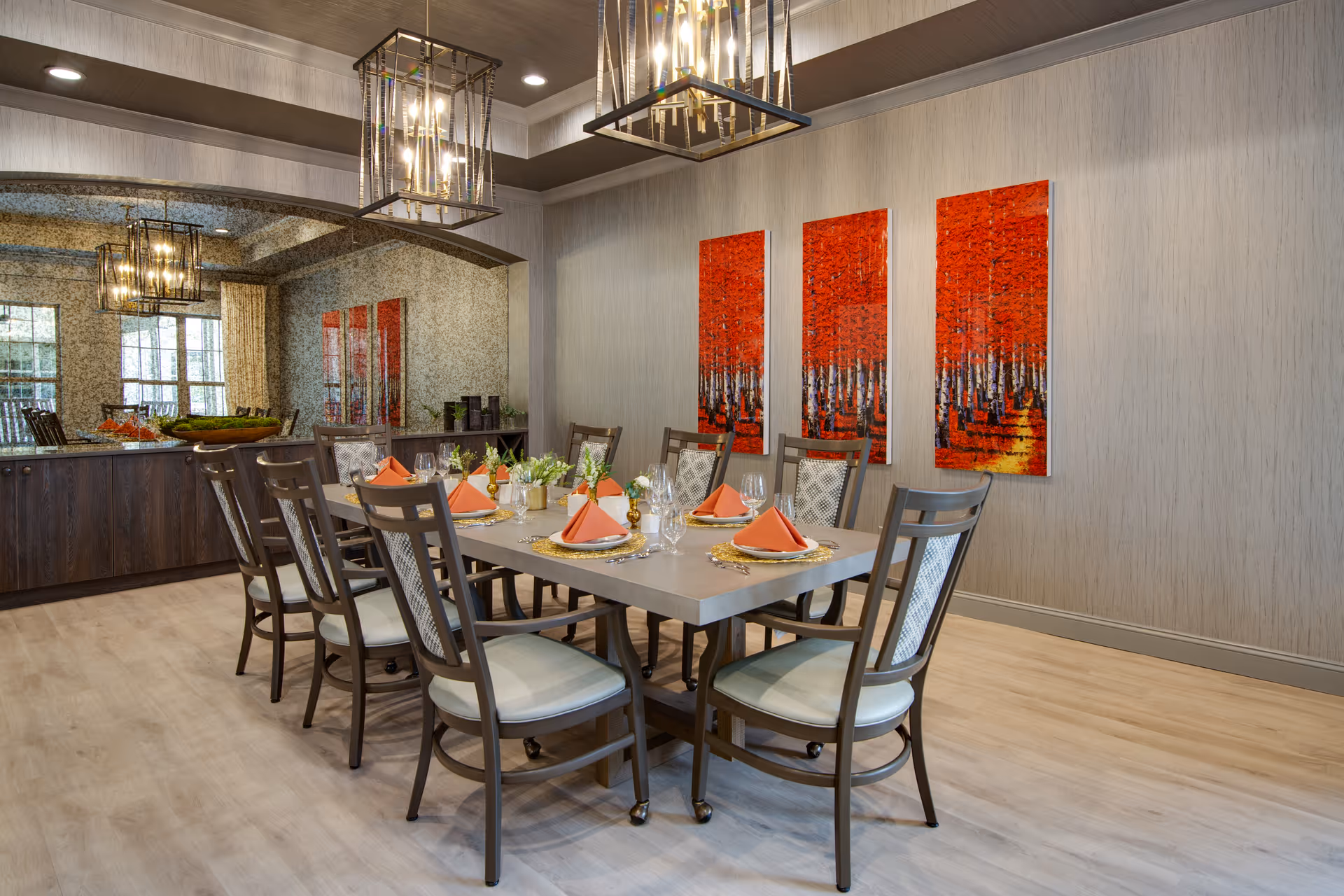 A modern dining room with a rectangular table set for eight people. The table is decorated with orange napkins, glassware, and small floral centerpieces. Behind the table, there are three vertical paintings featuring a forest with red foliage. The room has light wood flooring, a large mirror on one wall, and two rectangular pendant lights hanging from the ceiling.