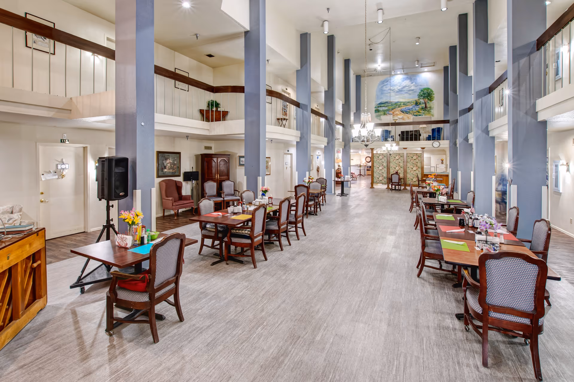 A spacious dining room in an independent living facility with multiple wooden tables and chairs arranged neatly. The room features high ceilings with tall vertical beams, chandeliers, and a large mural painting on the far wall. There are decorative plants and flowers on the tables, and a piano is visible on the left side. The area has a warm and inviting atmosphere with a mix of classic and modern decor.