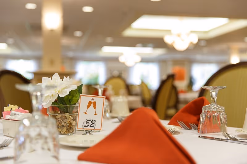 A dining table in a senior living dining room set with folded orange napkins, glassware, and a floral centerpiece with a table number.