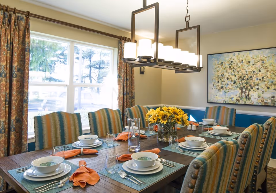 A dining room with a wooden table set for eight people. The table has blue placemats, white plates and bowls, silverware, clear drinking glasses, and orange cloth napkins. A vase with sunflowers is in the center of the table. The chairs have striped upholstery in shades of blue, green, and orange. There is a large window with patterned curtains letting in natural light, a modern rectangular chandelier with multiple lights hanging above the table, and a colorful abstract painting on the wall.