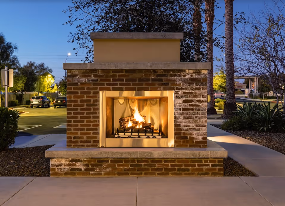 Outdoor brick fireplace with a fire burning inside, surrounded by a paved walkway, trees, and parked cars in the background during dusk.