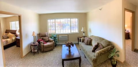 A cozy living room with a large window letting in natural light. The room features a green upholstered sofa with several pillows, a reclining chair with a red blanket draped over one arm, a wooden coffee table with a flower arrangement, two side tables with lamps, and a potted plant near the sofa. Doorways on either side lead to bedrooms.