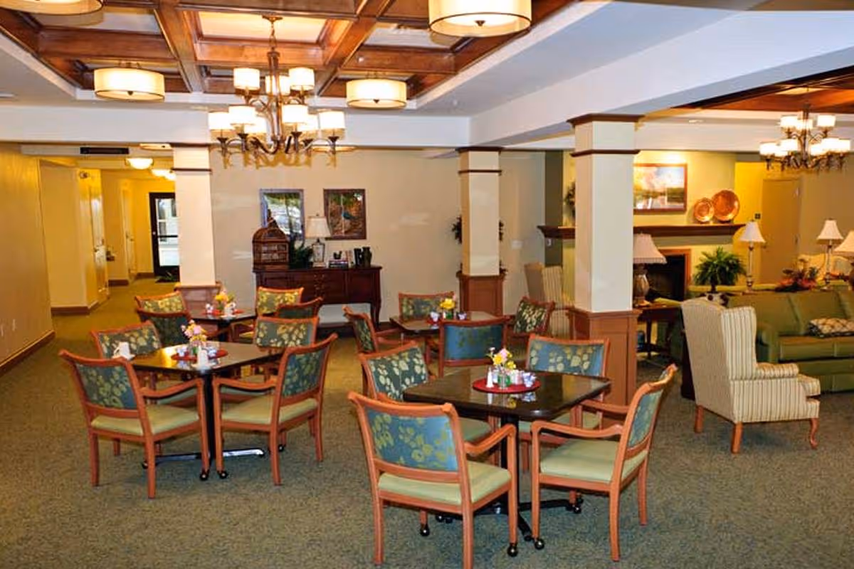 A cozy common area in a senior living facility with multiple small tables and chairs arranged for socializing or dining. The room features a green carpet, wooden ceiling beams with light fixtures, and a seating area with sofas and armchairs near a fireplace. Decorative plants and framed artwork adorn the walls.