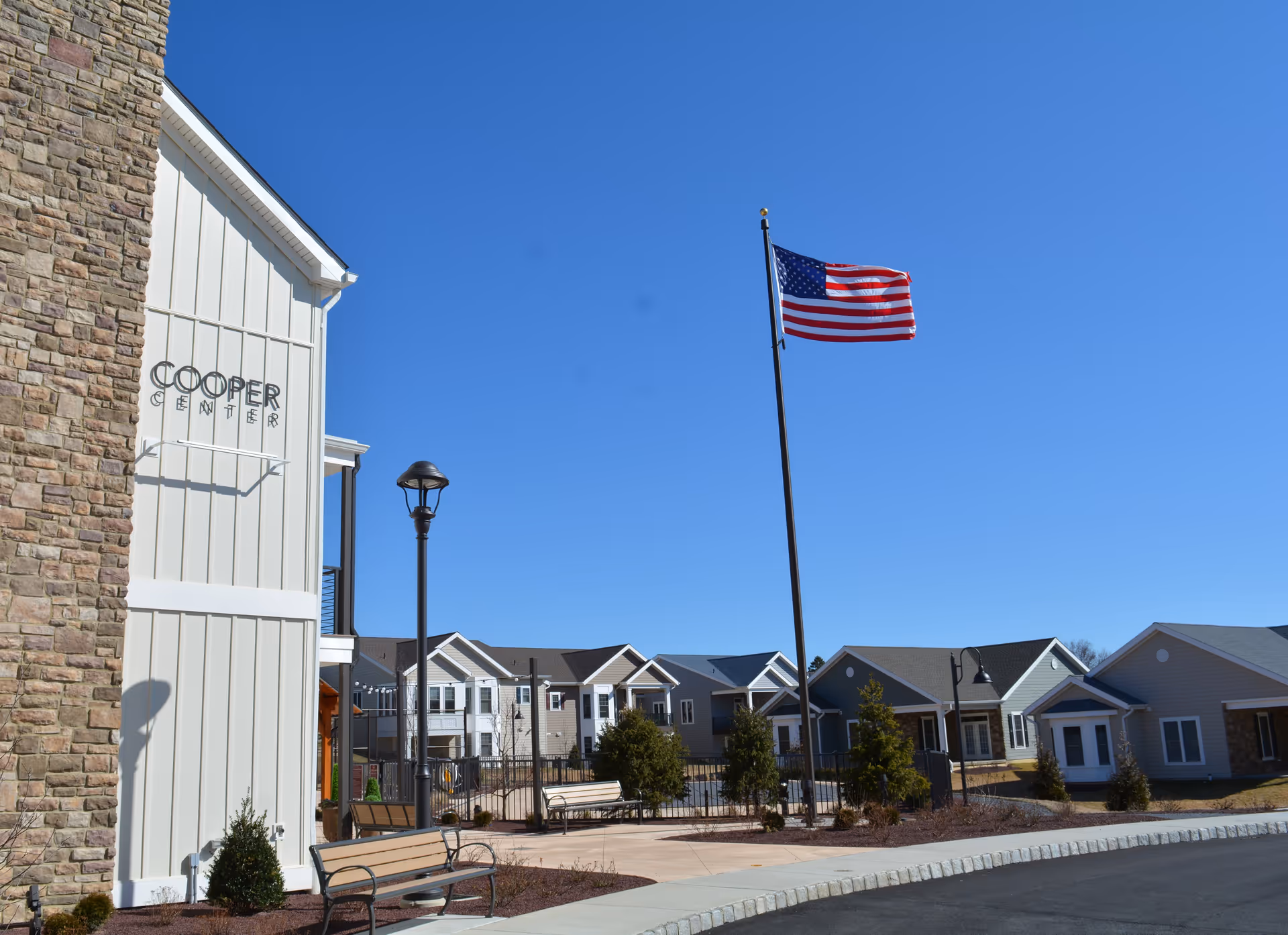 Outdoor view of a senior living facility named Heritage Village showing the Cooper Center building on the left, a tall flagpole with an American flag waving, benches, street lamps, and several single-story residential buildings under a clear blue sky.