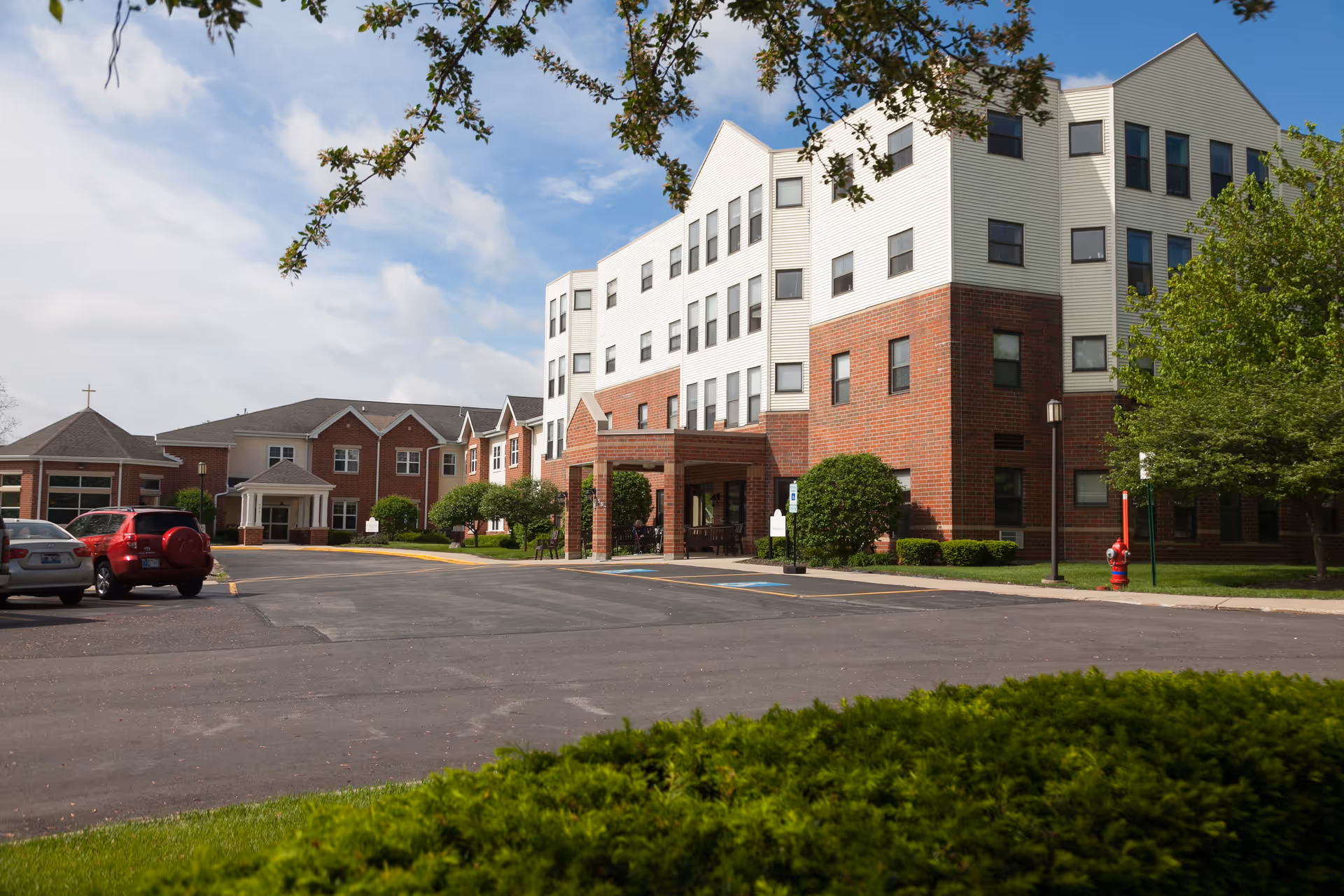 Exterior view of Addolorata Villa, a multi-story senior living facility with a combination of red brick and white siding. The building has multiple windows and a covered entrance. There are a few cars parked in the parking lot, green bushes, and trees surrounding the area under a partly cloudy sky.