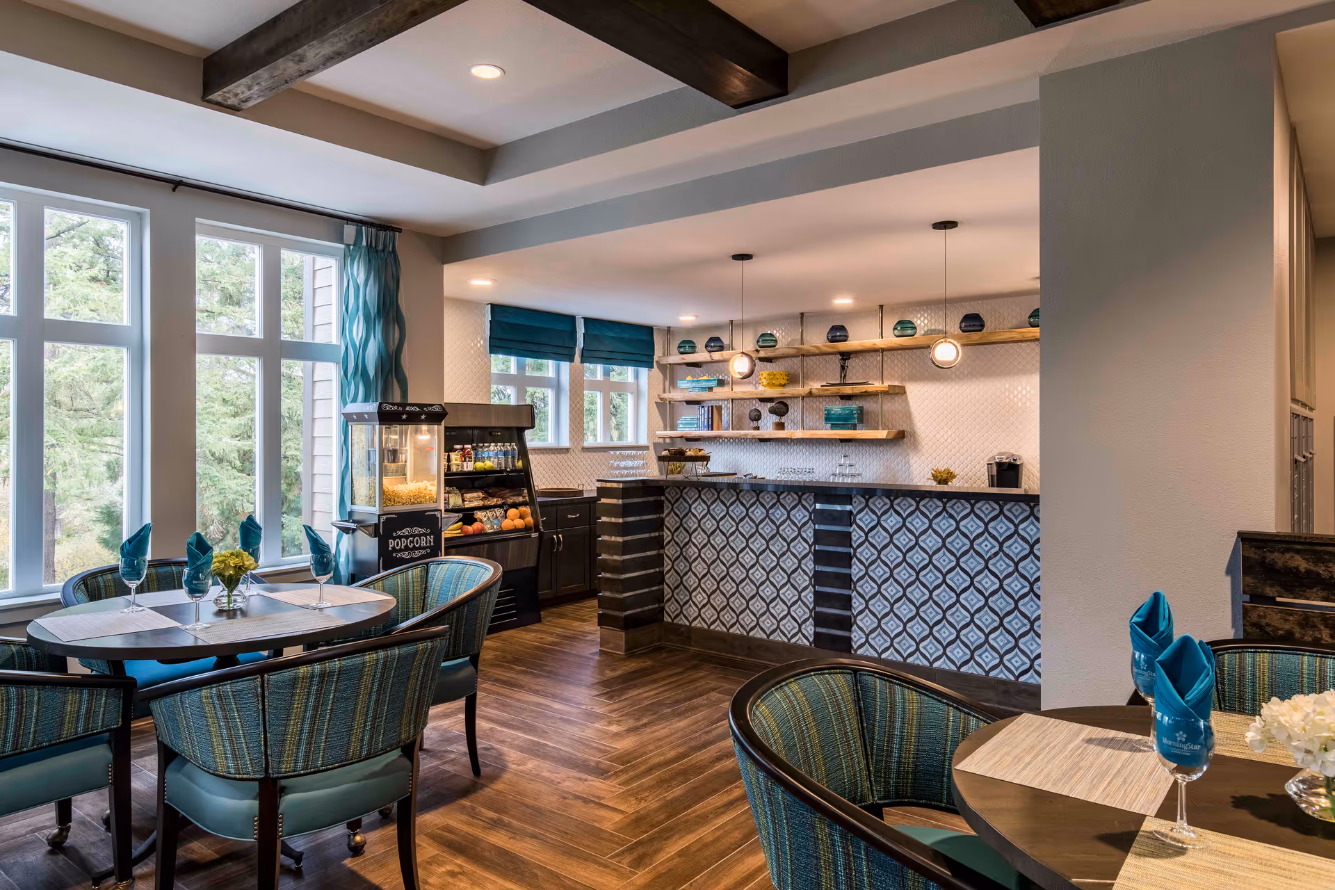 A bright dining area in a senior living facility with round tables and teal cushioned chairs. Each table is set with placemats, folded teal napkins in glasses, and small flower arrangements. Large windows with teal curtains allow natural light to fill the room. In the background, there is a counter with patterned tiles and shelves holding decorative items. A popcorn machine and a refrigerated display case with snacks and drinks are also visible.