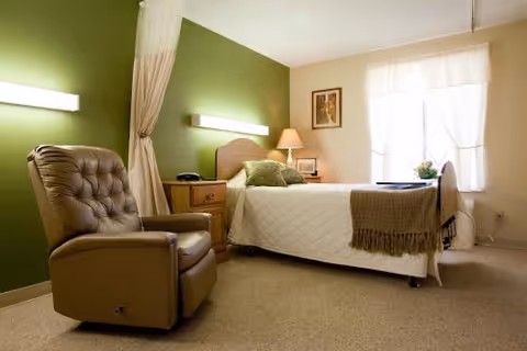 A tidy senior resident bedroom with a single bed, bedside table and lamp, and a recliner chair against a green accent wall.