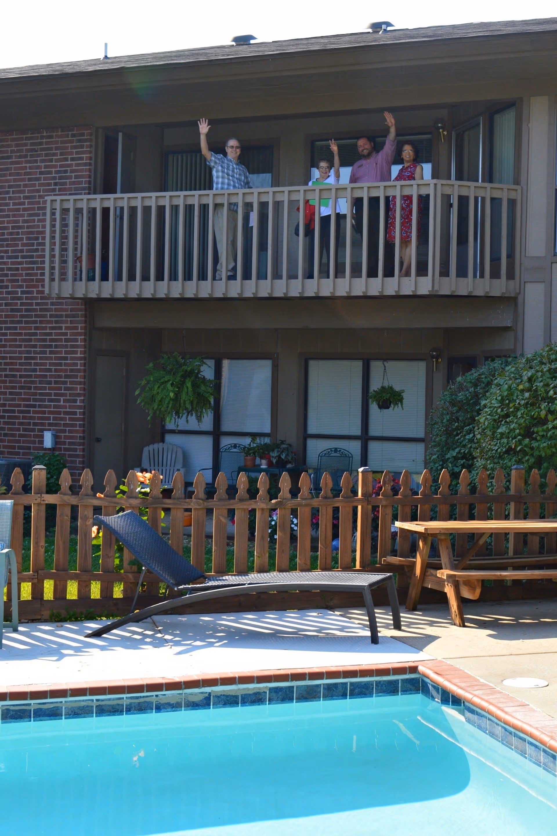 Four people standing on a second-floor balcony of a brick and wood building, waving. Below the balcony is a fenced garden area with hanging plants, chairs, and a wooden picnic table. In the foreground, there is a swimming pool with clear blue water and a black lounge chair on the pool deck.