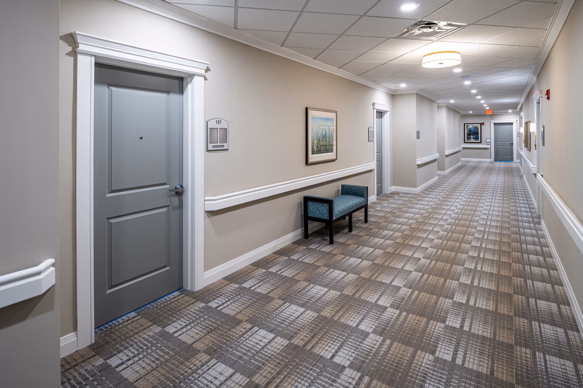 A long, well-lit hallway in a senior living facility with beige walls, patterned carpet flooring, and several closed gray doors. There is a small bench with blue upholstery along the wall and framed artwork hanging above it. The ceiling has recessed lighting and a circular light fixture.