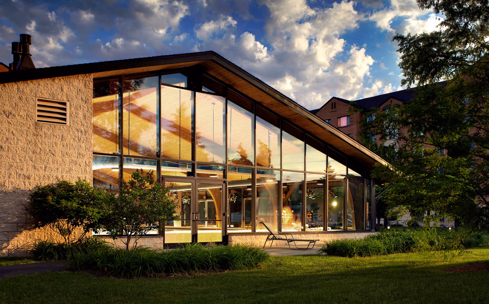Exterior view of a modern building with large glass windows reflecting the sky and surrounding trees, set against a partly cloudy sky during sunset. The building is surrounded by green grass, bushes, and trees, with a lounge chair visible near the windows.