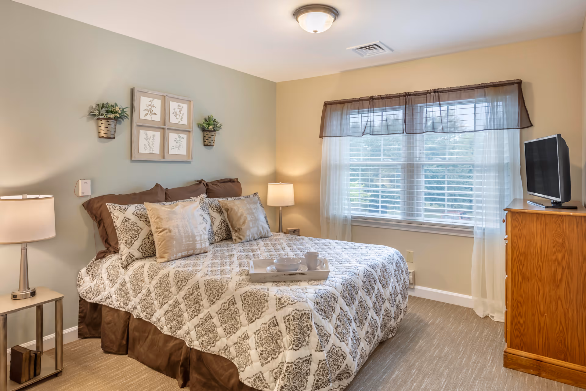 A cozy bedroom with a large bed covered in a patterned quilt and multiple pillows. There are two bedside tables with lamps on either side of the bed. Above the bed, there are four framed botanical prints and two small wall-mounted plant holders with greenery. A window with sheer curtains and a dark valance lets in natural light. A wooden dresser with a flat-screen TV on top is positioned near the window.