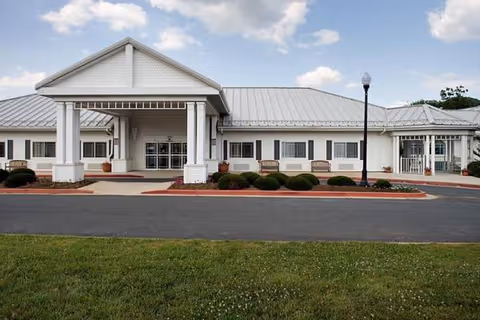 Front exterior of a senior living facility with a covered entrance, benches, and manicured shrubs.