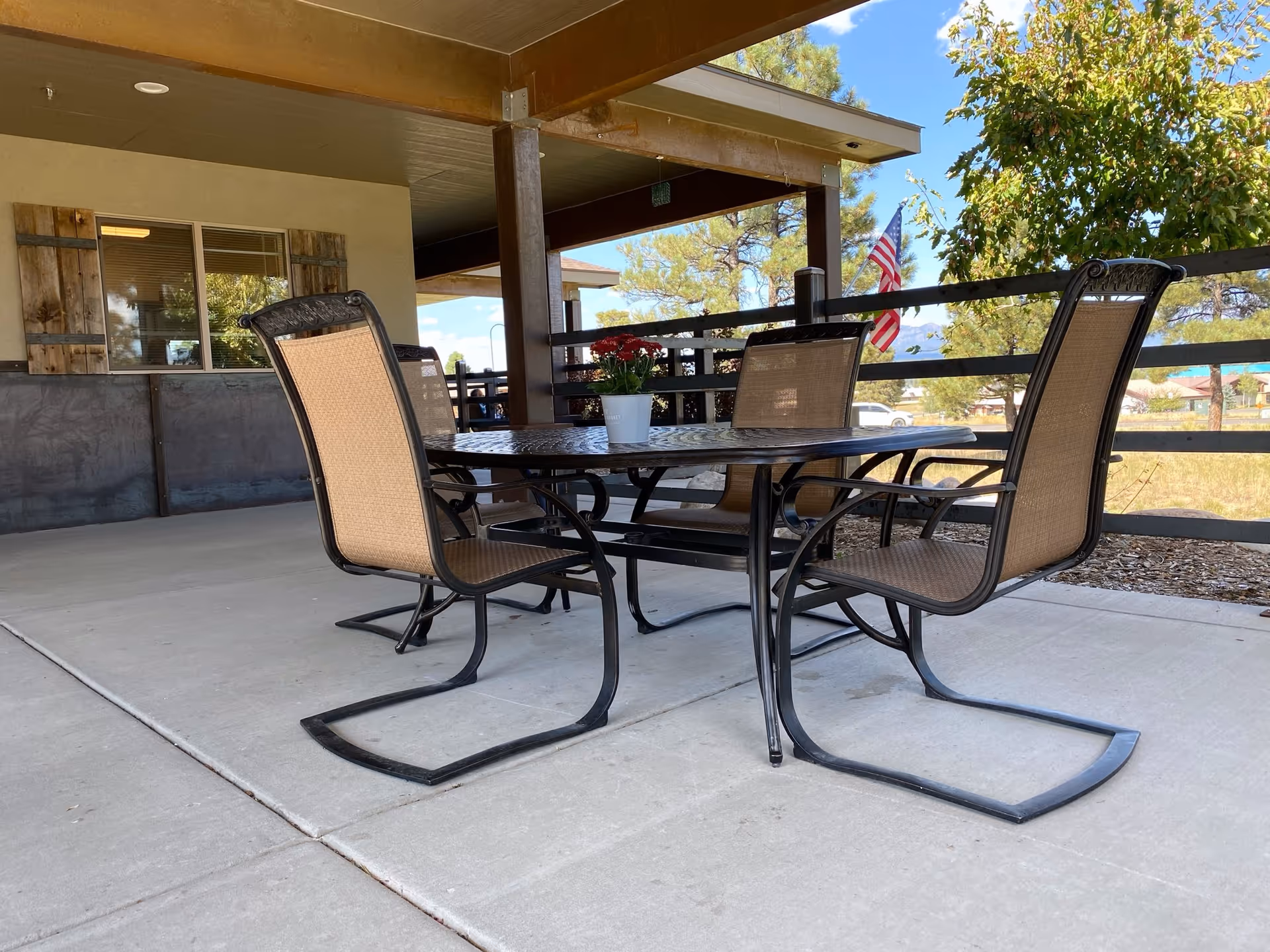 Outdoor patio area with a metal table and four mesh chairs on a concrete floor. A small potted plant with red flowers is placed on the table. The patio is covered with a roof and has wooden posts and railing. Trees, an American flag, and a clear blue sky are visible in the background.