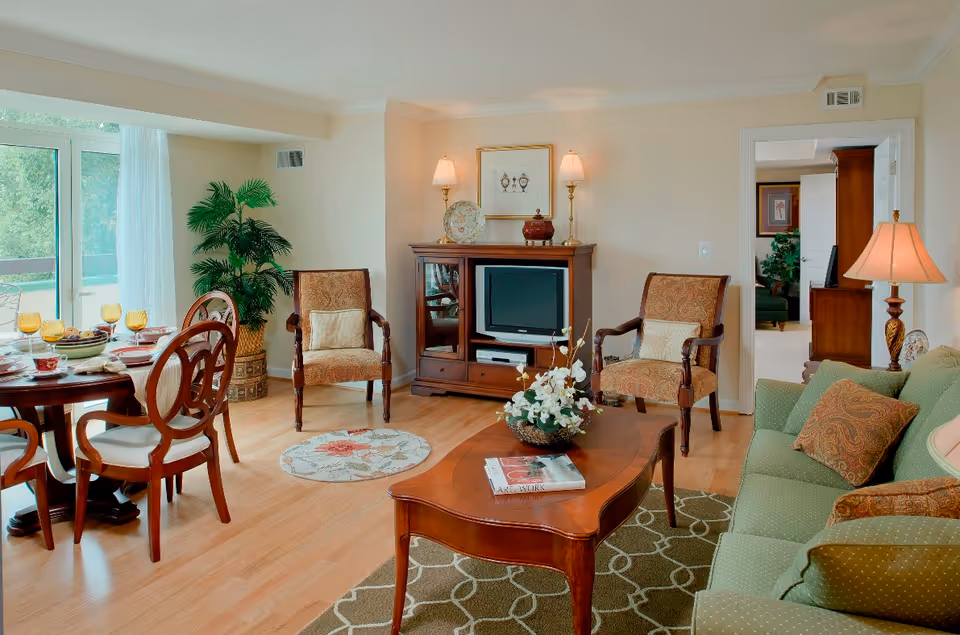 A cozy living room with a green sofa adorned with patterned cushions, two upholstered armchairs, a wooden coffee table with a floral centerpiece and a book, and a wooden TV cabinet with a small television. To the left, there is a dining area with a round wooden table set with plates, glasses, and napkins. A large window with sheer white curtains lets in natural light, and a potted plant is placed near the corner. The room has light-colored walls and hardwood flooring, with a patterned rug under the coffee table.