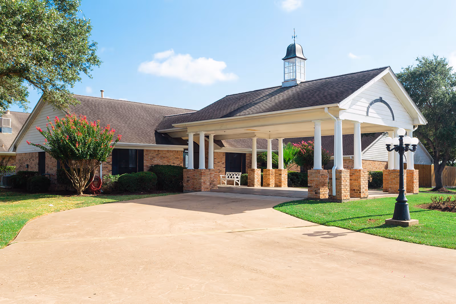 Exterior view of a single-story brick building with a covered entrance supported by white columns and brick bases. There is a concrete driveway leading up to the entrance, green grass, a black lamp post with multiple white globes, and trees and shrubs surrounding the building under a blue sky with some clouds.