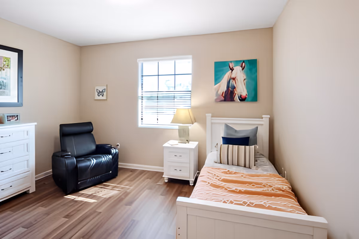 A cozy bedroom with a single white bed covered with an orange blanket and multiple pillows. Next to the bed is a white nightstand with a lamp. A black leather recliner chair is positioned near a white dresser. The room has beige walls, a window with blinds, and two framed pictures on the walls, one of a horse and one of a butterfly.