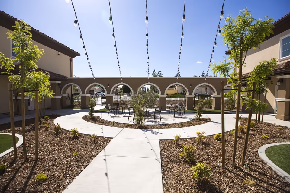 Outdoor courtyard area with a circular paved walkway and several small trees and plants. There are string lights hanging overhead and several tables with chairs arranged in the center. The courtyard is surrounded by beige buildings with arched openings and a clear blue sky above.