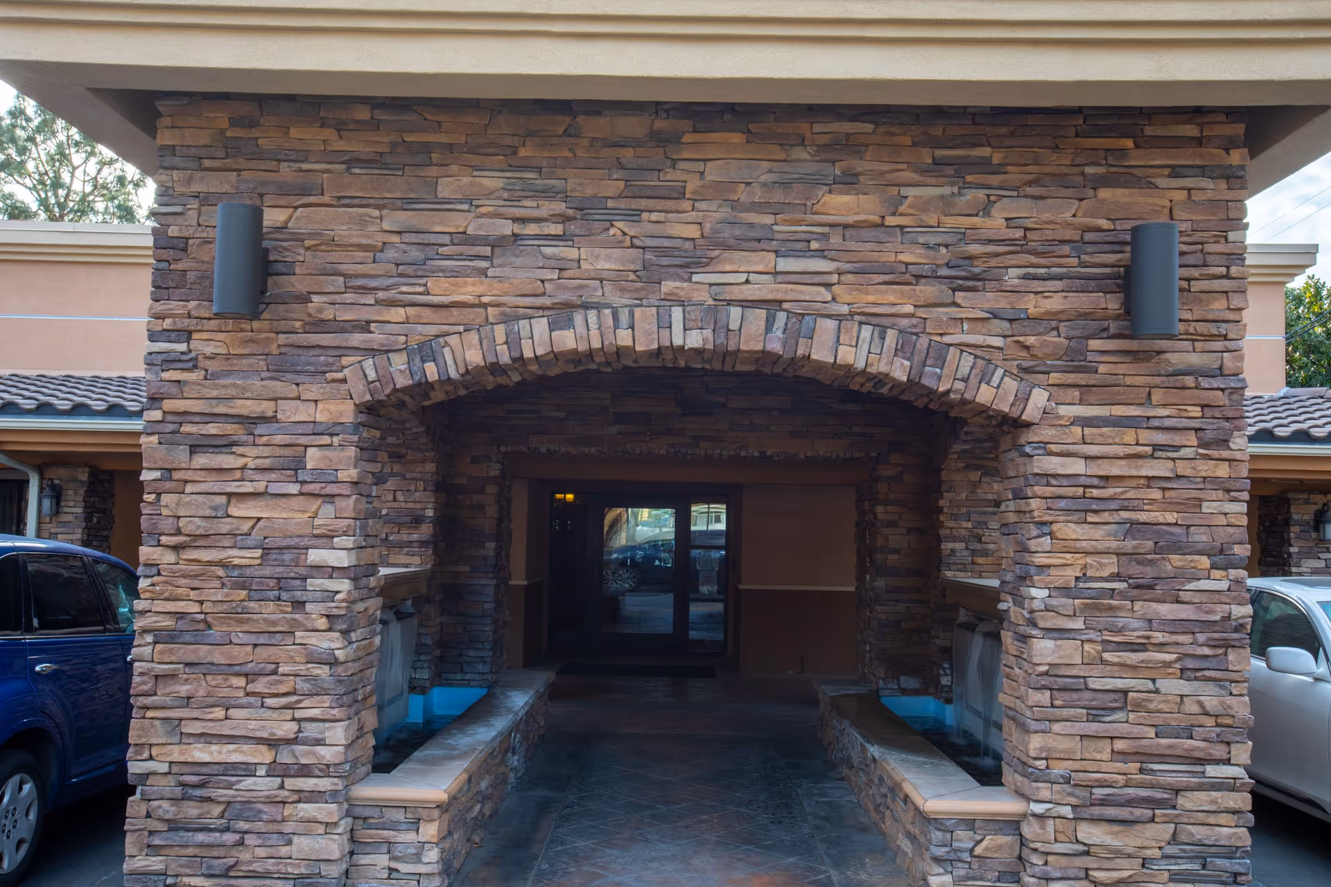 Stone archway entrance to a building with two water features on either side and parked cars visible on both sides.