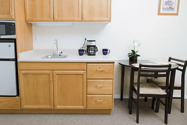 Small kitchenette area with wooden cabinets, a countertop with a sink, a coffee maker, and two blue mugs. To the right, there is a small round dining table with two chairs, a potted orchid plant, and a newspaper on the table. A framed picture hangs on the wall above the table.