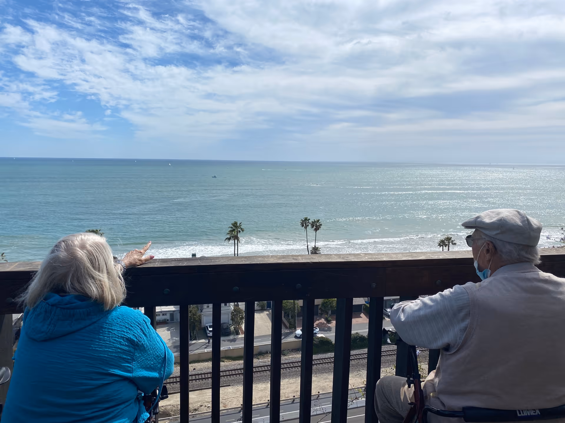 Two elderly individuals sitting on a balcony overlooking the ocean. One person in a blue jacket is pointing towards the sea, while the other person wearing a light-colored cap and mask looks on. The view includes palm trees, a railway track, and a partly cloudy sky.