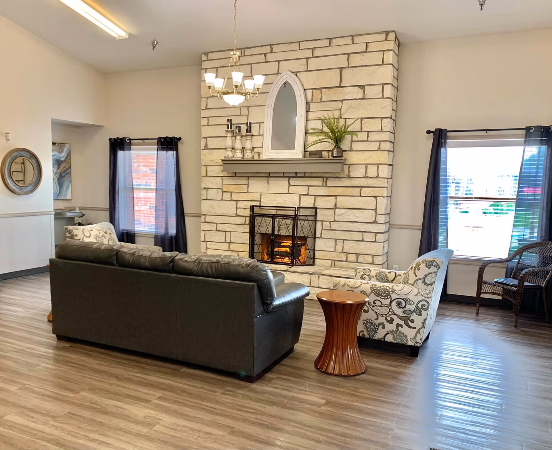A cozy living room area with a stone fireplace featuring a lit fire, a dark leather sofa, a patterned armchair, a small wooden side table, and two windows with dark curtains. The room has light-colored walls, wood flooring, and a chandelier hanging from the ceiling.