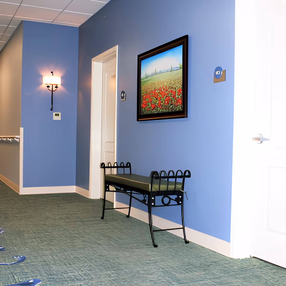 A hallway in a senior living facility with blue walls and green carpet. There is a black metal bench with a green cushion against the wall, a framed painting of a field with red flowers hanging above it, a wall-mounted light fixture, and a white door partially visible on the right side.