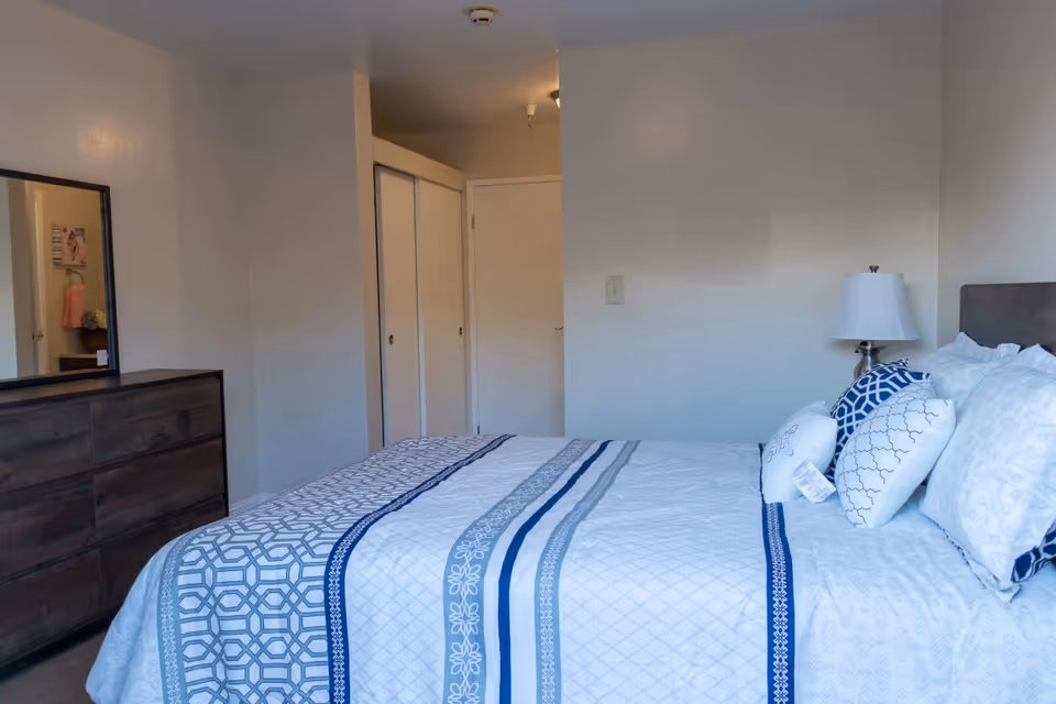 A simple bedroom with a neatly made blue-and-white bed, decorative pillows, a bedside lamp, dresser with mirror, and closet doors.