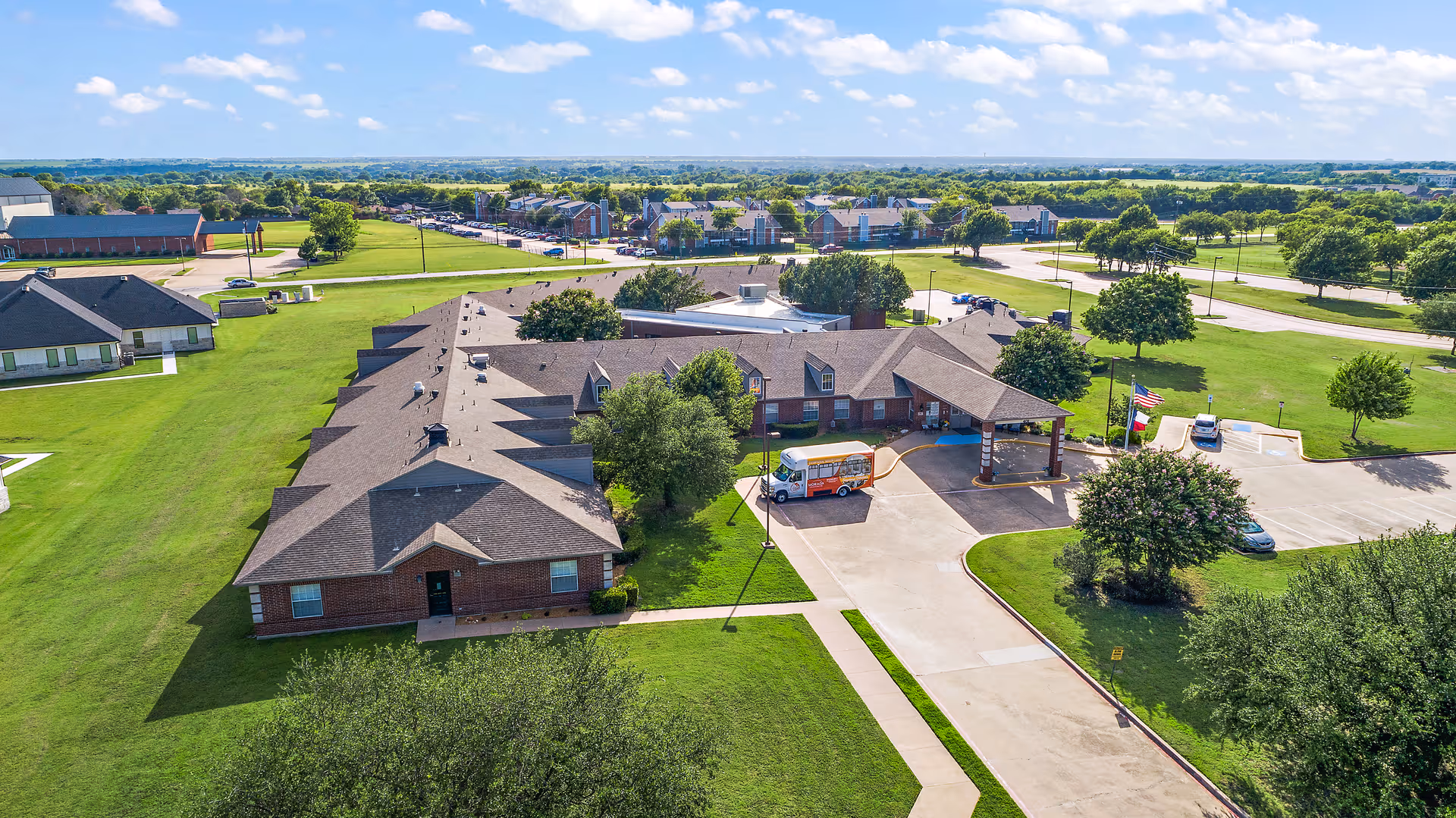 Aerial view of Morada Waxahachie senior living facility showing a large single-story brick building with a covered entrance, surrounded by green lawns, trees, and a parking area with a shuttle bus parked near the entrance. The sky is partly cloudy with a wide horizon in the background.