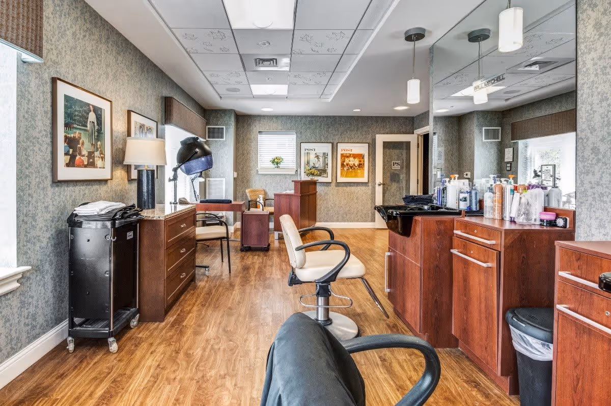 Interior view of a salon area in a senior living facility with wooden flooring, salon chairs, a hair dryer, cabinets with various hair care products, framed pictures on the walls, and large mirrors.