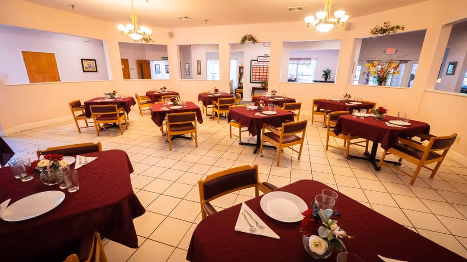A dining room with multiple square tables covered with burgundy tablecloths, each set with white plates, glasses, silverware, and small floral centerpieces. Wooden chairs with padded seats surround the tables. The room has tiled floors, light-colored walls, and several windows and openings to adjacent rooms.