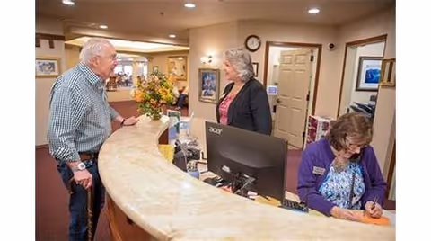 An elderly man with a cane is standing at a curved reception desk talking to a woman behind the desk, while another woman is seated and writing at the same desk in a well-lit interior space with beige walls and framed pictures.