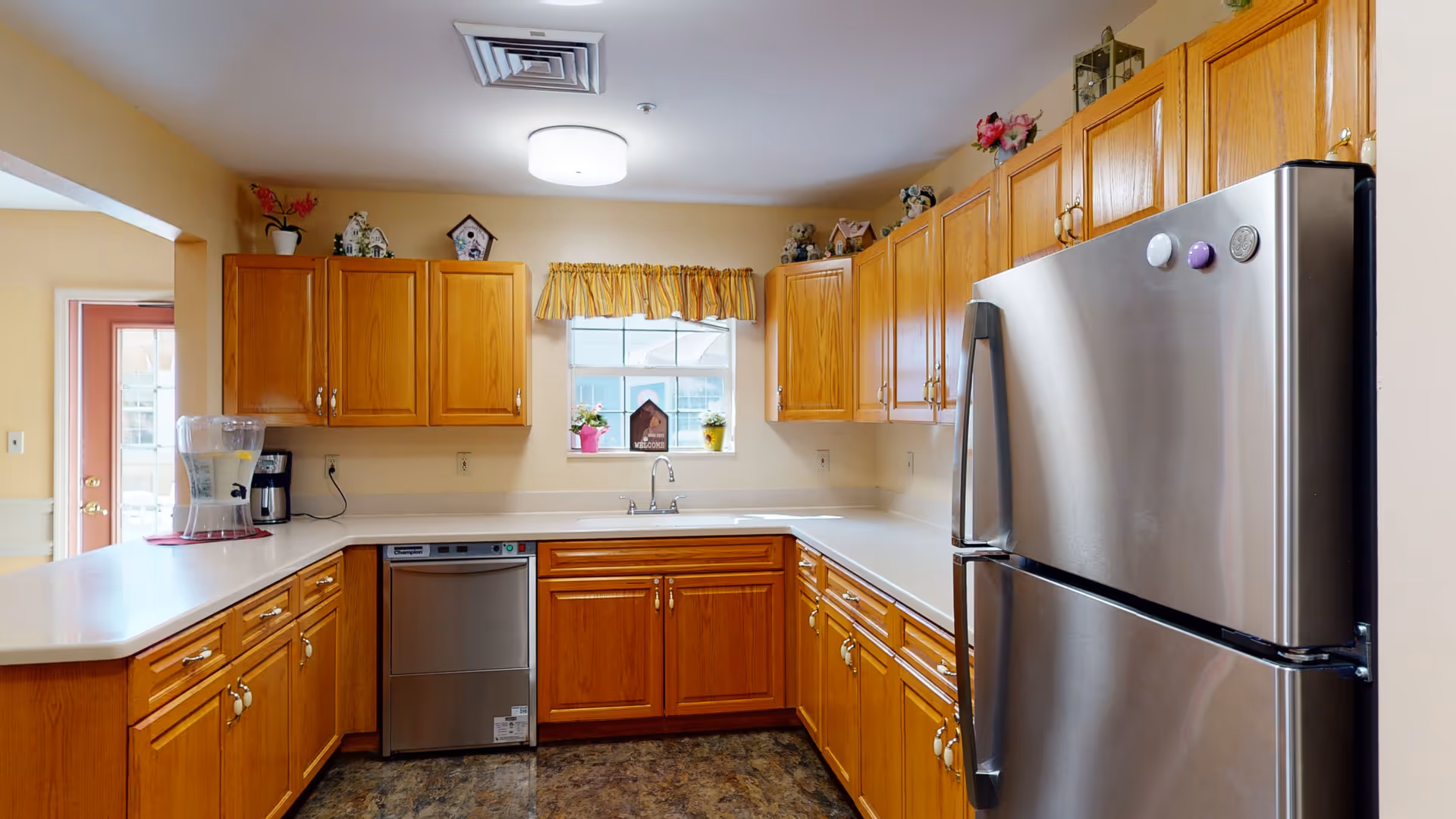 A clean and well-lit kitchen with wooden cabinets, a stainless steel refrigerator, a dishwasher, a coffee maker, and a water dispenser. There is a window with a yellow valance above the sink, and decorative items are placed on top of the cabinets.