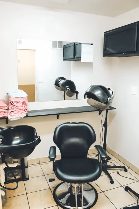 Interior of a small hair salon area with a black salon chair in front of a large mirror. There is a black hair washing sink to the left, a hair dryer hood on a stand to the right, and a shelf with folded pink towels. The walls are light-colored and the floor is tiled.
