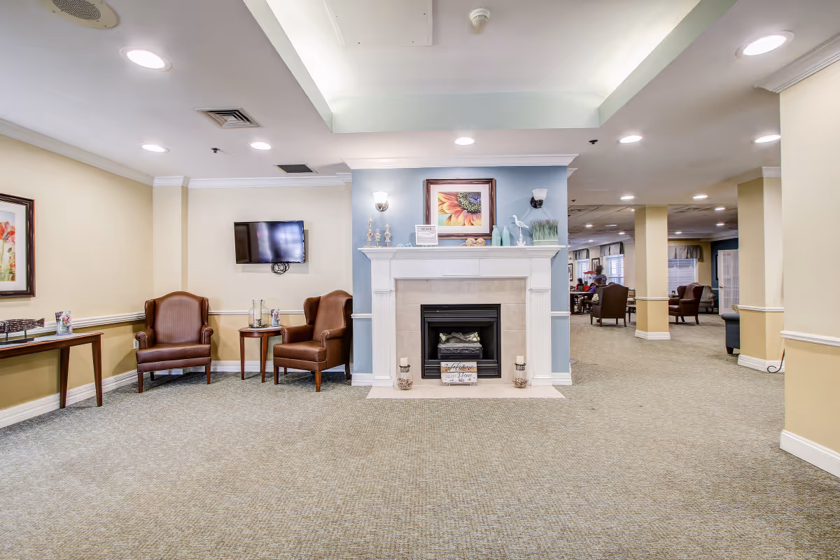 Well-lit senior living common area with a central fireplace, armchairs, a wall-mounted TV, and additional seating in a carpeted room.