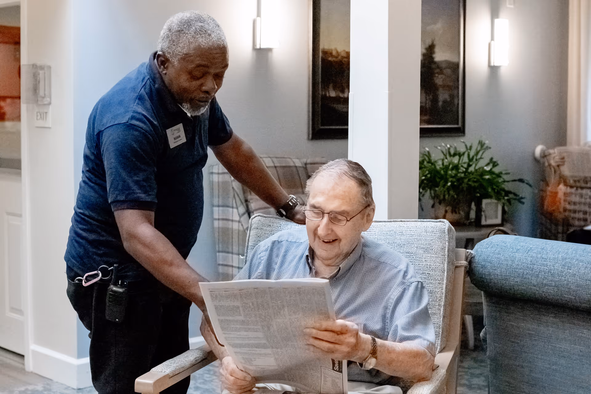 An elderly man sitting in a comfortable chair reading a newspaper while a staff member stands beside him, engaging and assisting in a well-lit living room with plants and framed artwork on the walls.