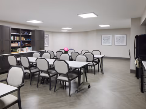 A well-lit interior room with multiple tables and chairs arranged in rows, a cabinet with books and decorative items on the left wall, two framed artworks on the far wall, and a refrigerator on the right side.