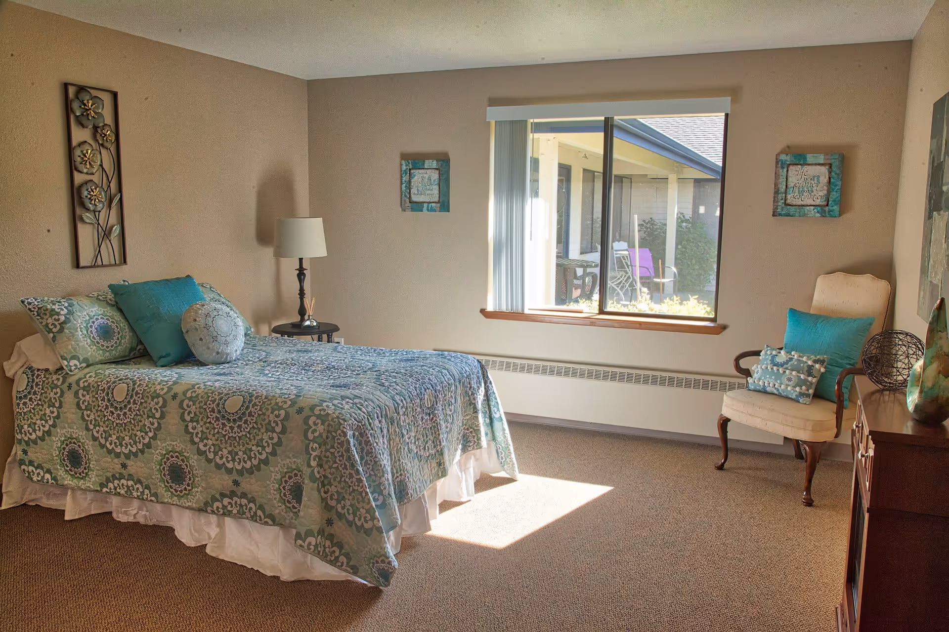 A cozy bedroom in a retirement community with a bed covered in a patterned blue and green quilt, matching pillows, a side table with a lamp, a window letting in natural light, a cushioned chair with blue pillows, and decorative wall art.