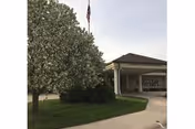 Exterior view of a building entrance with a covered driveway, a large tree with white blossoms, green bushes, and an American flag on a flagpole against a clear sky.