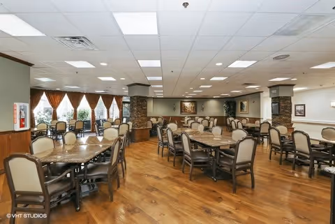 Spacious dining room with multiple tables and chairs arranged neatly on a wooden floor. Large windows with curtains allow natural light to brighten the room. The ceiling has recessed lighting and the walls are decorated with framed artwork and stone pillars.