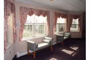 Sunlit seating area with three upholstered benches along a windowed wall and floral valances in a carpeted common room.