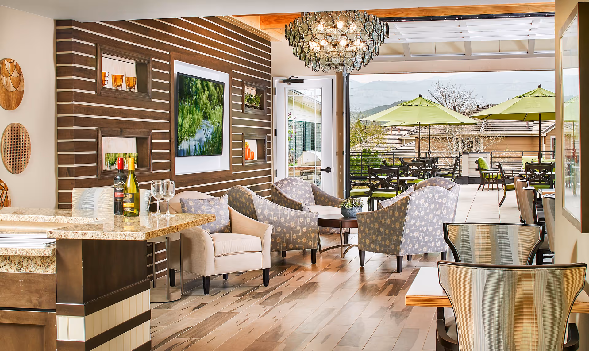 Bright communal lounge with armchairs, a granite-topped bar, a TV on a wood-paneled wall, and doors opening to a patio with green umbrellas.
