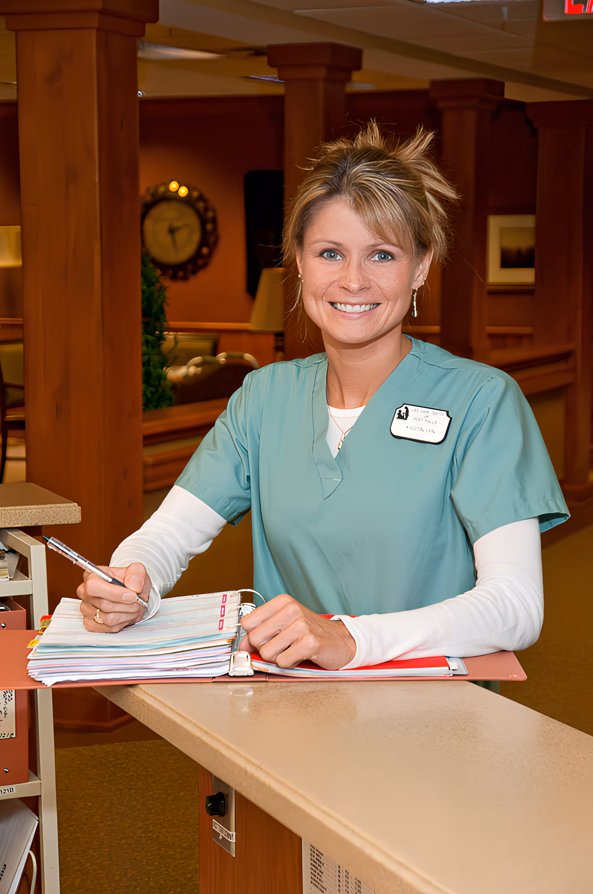 A smiling female healthcare worker in green scrubs and a white long-sleeve shirt is standing behind a counter, writing in a large binder. The background shows a warm-toned interior with wooden columns, a clock on the wall, and some seating areas.
