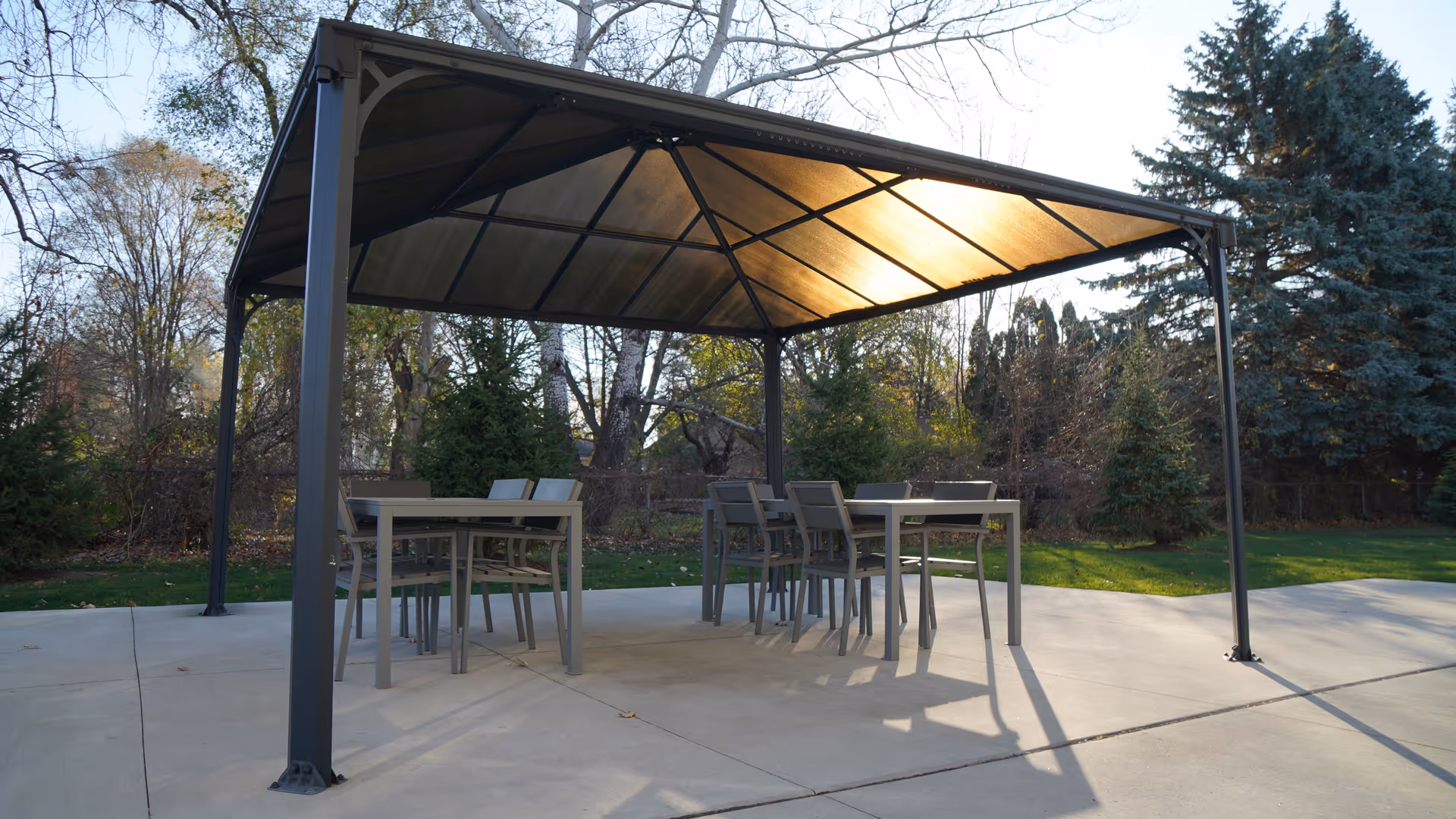 Outdoor patio area with a metal-framed gazebo providing shade over two tables and several chairs, surrounded by trees and greenery in the background.