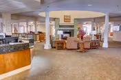 Spacious senior living facility common lounge with seating arranged around a central fireplace, columns, a reception desk, and windows.