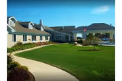 Exterior view of a single-story senior living facility building with beige siding and multiple dormer windows. A well-maintained green lawn and a curved concrete walkway lead to the entrance, which is covered by a large portico. The sky is clear and blue.